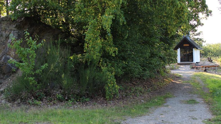 Ein schmaler Weg führt zu einer kleinen, weißen Hütte, umgeben von üppigem Grün. Die Szene strahlt Ruhe und Naturverbundenheit aus.