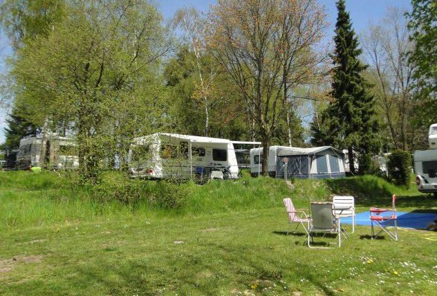 Ein Campingplatz mit Wohnmobilen und einem Zelt in einer grünen Umgebung. Im Vordergrund stehen zwei Stühle auf der Wiese.