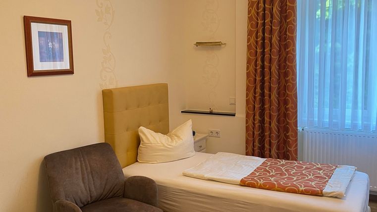 Ein einfaches, gemütliches Schlafzimmer mit einem Einzelbett und einem Stuhl. Große Fenster lassen viel Tageslicht herein.