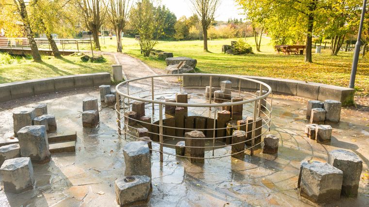 Ein Wasserspielplatz mit runden Steinen und einer metallenen Umrandung. Umgeben von Bäumen und grünem Gras.