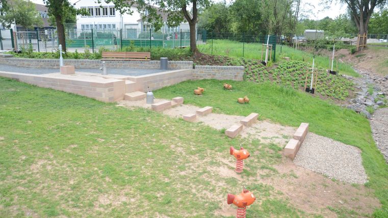 Een speelplaats met grasvelden en verschillende speeltoestellen. Op de achtergrond zijn bomen en een klein stuk met grind te zien.