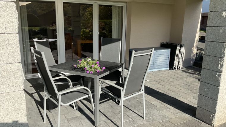 A cozy outdoor area with a table and four chairs. A flower bowl is on the table, and large windows can be seen in the background.