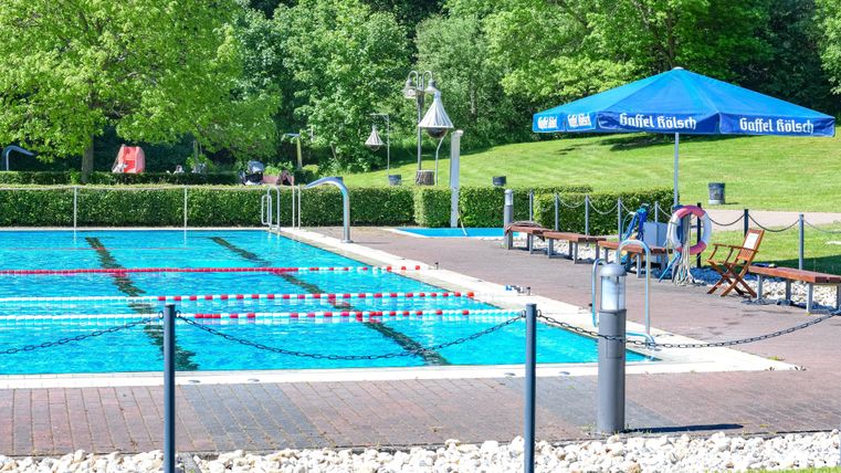 Een zwembad met helder water, omringd door groene bomen. Langs de rand van het bad staan ligstoelen en een parasol.