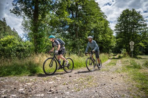 Two cyclists are riding on a gravel path in a green woodland area. The sky is partly cloudy and the surroundings appear inviting and natural.