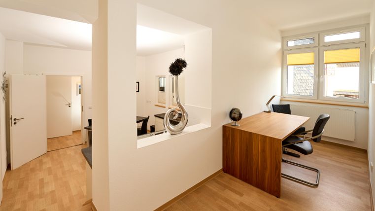 Ein modern eingerichtetes Büro mit Holzmöbeln und viel Tageslicht. Es gibt einen Schreibtisch, einen Stuhl und eine dekorative Pflanze.