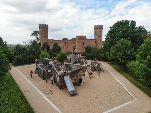 Ein Spielplatz mit einer Burgenstruktur und Rutschen, umgeben von Bäumen. Im Hintergrund sieht man eine beeindruckende Burg mit Türmen.