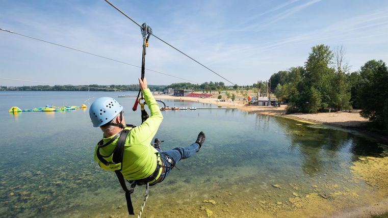 Eine Person fährt mit einer Zipline über einen klaren See. Im Hintergrund sind Bäume und ein Strand zu sehen.
