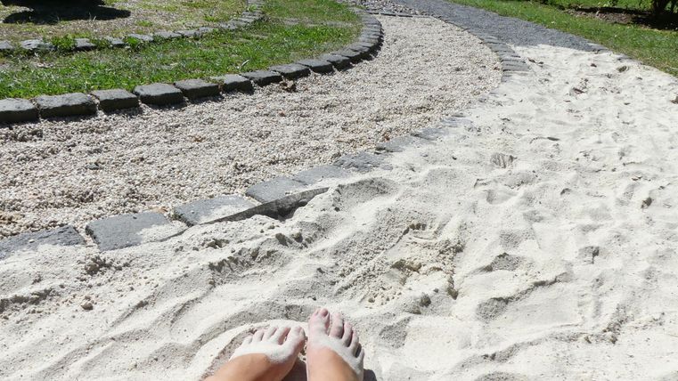 Ein schmaler Sandweg in einem grünen Garten. Im Vordergrund sind Füße im Sand zu sehen.