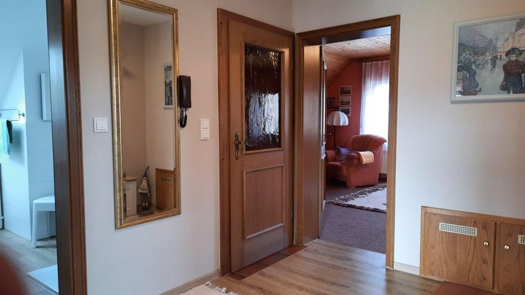 A bright hallway with a mirror and a wooden interior door. In the background, a cozy living room with a red sofa is visible.