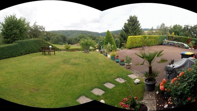 Ein großer Garten mit Grünflächen und vielen Pflanzen. Im Hintergrund sind Hügel und Bäume zu sehen, der Himmel ist bewölkt.