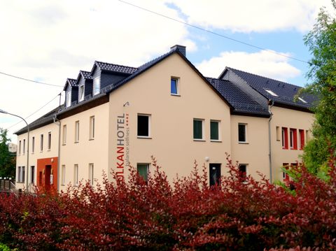 Ein modernes Hotelgebäude mit einem klaren, freundlichen Design. Im Vordergrund sind rotbunte Büsche zu sehen, die eine einladende Atmosphäre schaffen.