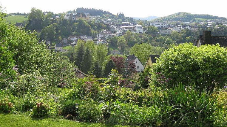 Een schilderachtig landschap met een mooie tuin op de voorgrond en een klein stadje op de achtergrond. De omgeving is groen en heuvelachtig, onder een heldere lucht.