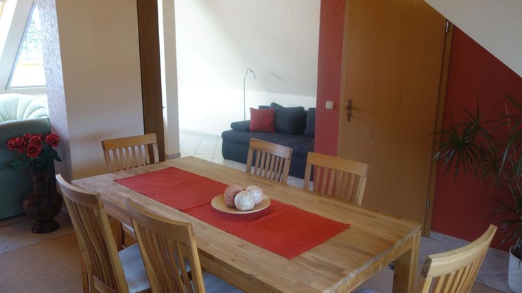 Een gezellige eetruimte met een houten tafel en stoelen. Op de achtergrond staat een grijze bank en een plantenarrangement.