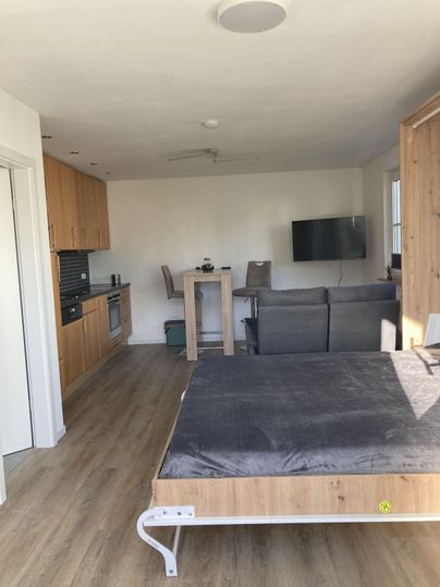 Eine moderne Wohnung mit einer offenen Küche und einem gemütlichen Wohnzimmer. Im Vordergrund befindet sich ein Bett mit einem grauen Bezug.