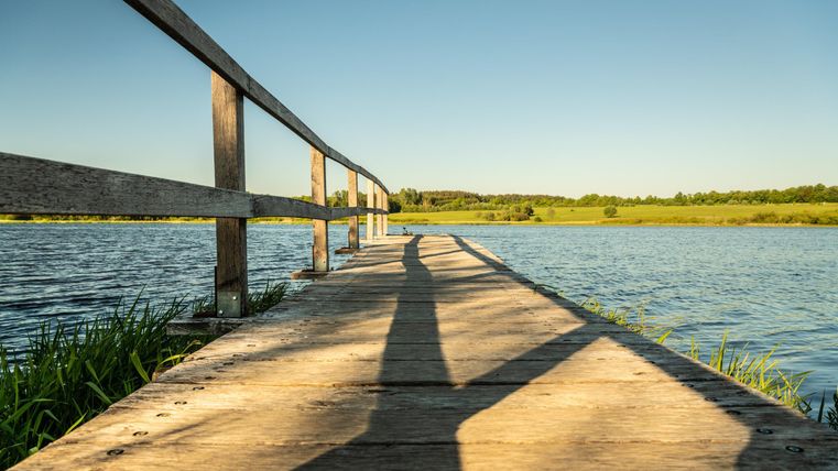 Ein Holzsteg führt über das Wasser in eine grüne Landschaft. Die klare Himmel und das ruhige Wasser schaffen eine friedliche Atmosphäre.