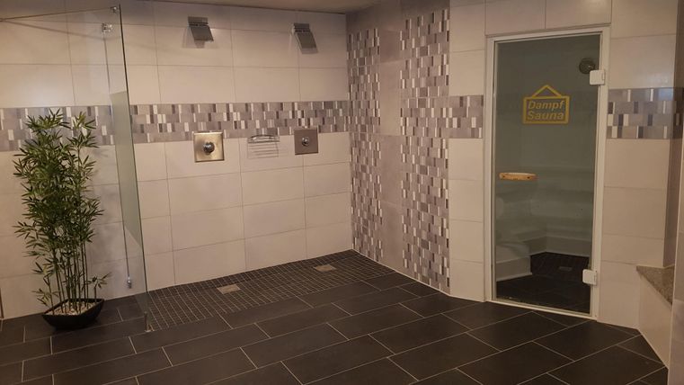 Ein modernes Spa mit weißen Fliesen und eleganten Akzenten. Auf der linken Seite steht eine Pflanze, und im Hintergrund ist eine Glasdusche sichtbar.