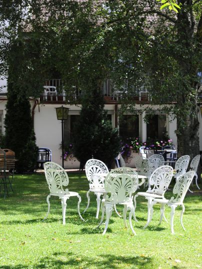 Ein schöner Garten mit weißen Gartenmöbeln. Bäume spenden Schatten und im Hintergrund sind Gebäude sichtbar.