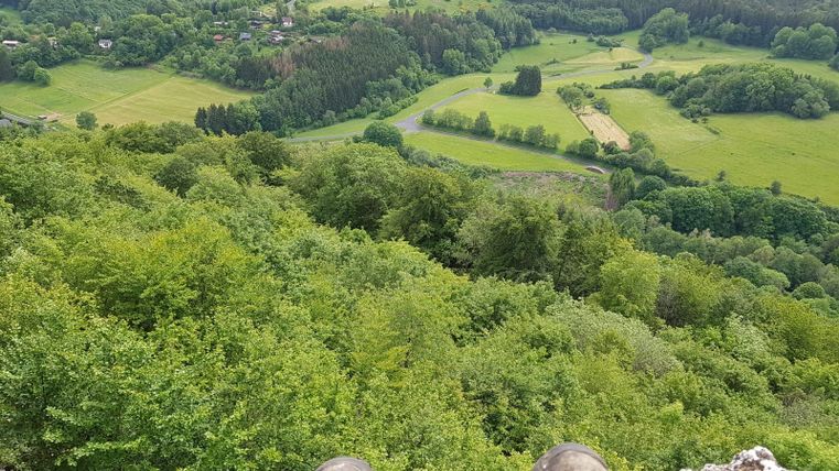 Een indrukwekkend uitzicht op een groene landschap met bossen en weiden. Op de voorgrond zijn laarzen op een steen zichtbaar.