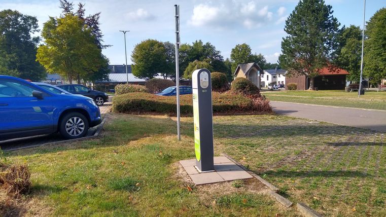 Eine Ladestation für Elektroautos steht auf einem Parkplatz. Im Hintergrund sind Bäume und ein ruhiger Wohnbereich zu sehen.