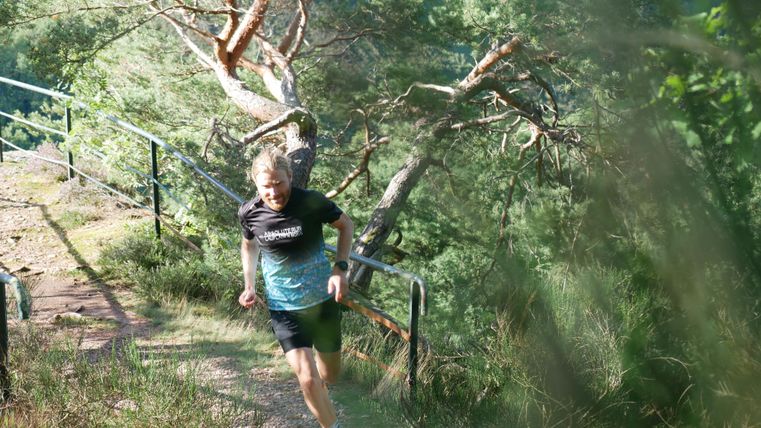 Ein Mann joggt auf einem schmalen Waldweg. Im Hintergrund sind Bäume und ein klarer Himmel zu sehen.