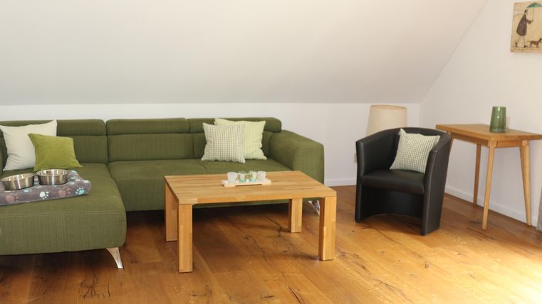 A cozy living area with a green couch and a wooden table. Next to it is a black armchair and a lamp on the wooden floor.
