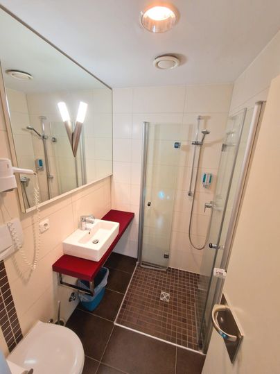 A modern bathroom with a shower cubicle and a sink on a red shelf. The walls are light tiled and the floor is dark tiled.