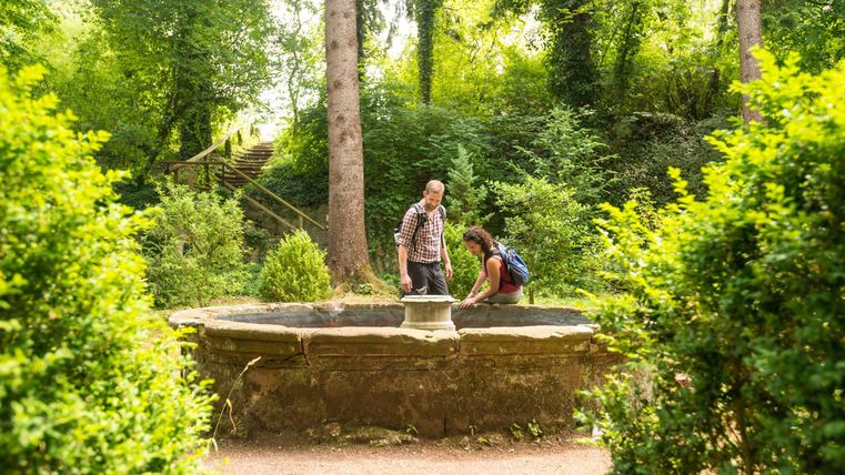 Ein Paar steht an einem alten Brunnen in einem grünen Wald. Umgeben von Bäumen wirken sie neugierig und entspannt.