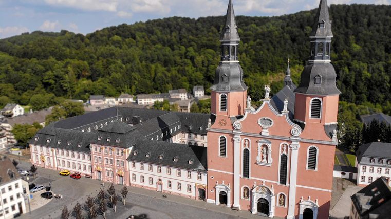 Eine beeindruckende Kirche mit zwei hohen Türmen steht im Mittelpunkt, umgeben von historischen Gebäuden und einem großen Platz. Im Hintergrund sind grüne Hügel zu sehen und der Himmel ist teilweise bewölkt.