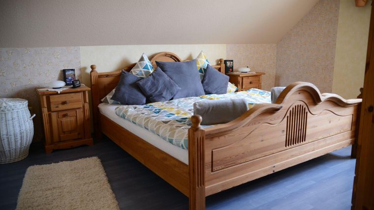 Een gezellige slaapkamer met een groot houten bed en kleurrijke kussens. Naast het bed staan twee nachtkastjes en een mand.