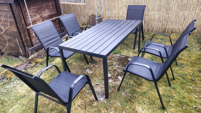 Ein Gartentisch mit sechs schwarzen Stühlen steht auf grünem Rasen. Im Hintergrund ist eine Holzwand sichtbar.