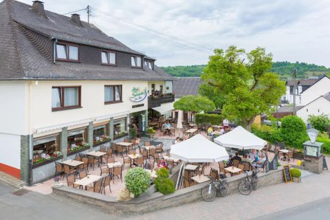 Ein gemütliches Café mit einer Terrasse und vielen Tischen im Freien. Im Hintergrund sind Bäume und eine ruhige Umgebung zu sehen.