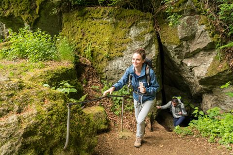 Zwei Wanderer kommen aus einer moosbewachsenen Höhle. Eine Frau vorne lächelt, während sie eine Treppe hinaufsteigt. Ein Mann folgt ihr.