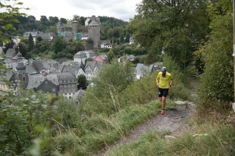 Ein Wanderer auf einem Pfad mit Blick auf eine malerische Stadt und eine Burg im Hintergrund. Die Umgebung ist grün und hügelig.