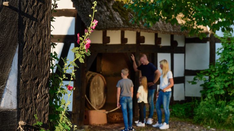 A family stands in front of a traditional half-timbered house and looks at a historic moat. In the foreground, flowers and trees can be seen.