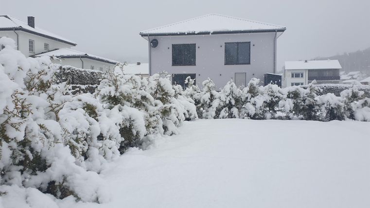 Ein verschneites Grundstück mit einem weißen Haus im Hintergrund. Die gesamte Landschaft ist mit einer dicken Schneedecke bedeckt.
