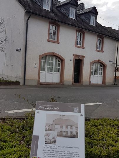 Ein historisches Gebäude mit einem hellen, schlichten Äußeren und einem Satteldach. Im Vordergrund steht ein Informationsschild über das Gebäude.