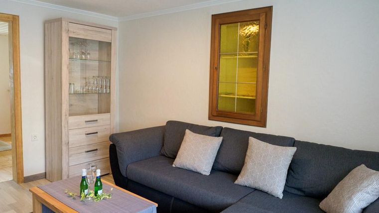 Ein gemütliches Wohnzimmer mit einem grauen Sofa und dekorativen Kissen. Ein Holztisch mit Wasserflaschen steht vor einer Glasvitrine.