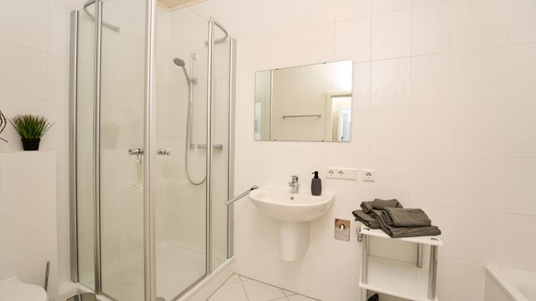 Ein modernes Badezimmer mit einer Dusche, einem Waschbecken und einem Spiegel. Weiße Wände und schlichte Ausstattung sorgen für ein sauberes, helles Ambiente.