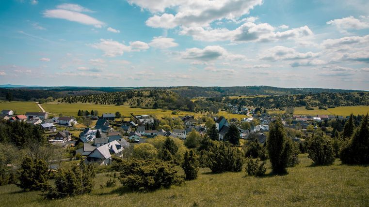 A picturesque view of a small village with green meadows and hills. The sky is clear and blue, with a few clouds.