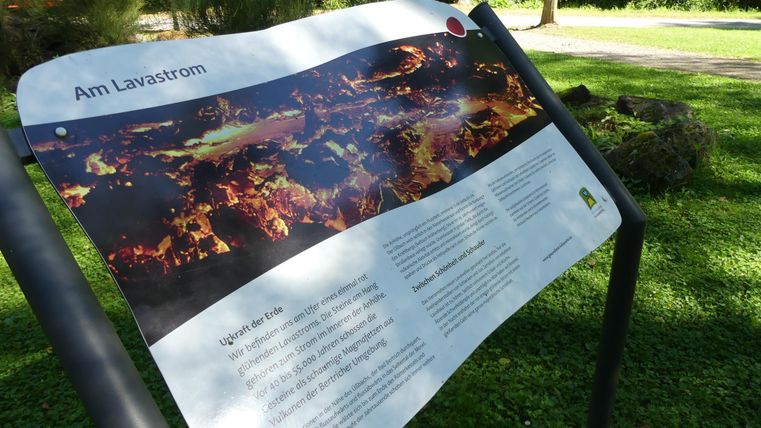 Ein Informationsschild über einen Lavastrom in einem Park. Im Hintergrund sind Bäume und grüne Wiese zu sehen.