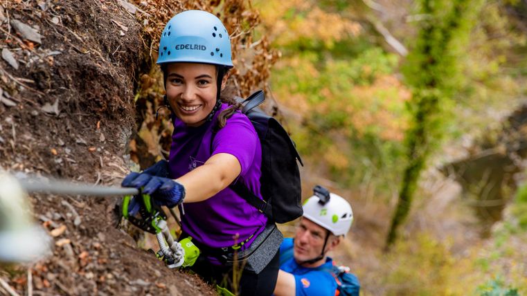 Eine Klettererin in einem lila Shirt und Helm steigt an einer Felswand. Im Hintergrund ist ein weiterer Kletterer zu sehen.