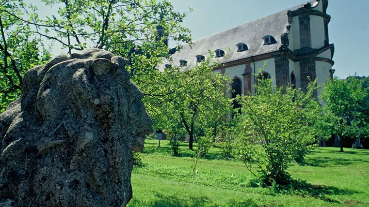 Eine alte Kirche hinter einem grünen Rasen und Obstbäumen. Im Vordergrund ist eine steinerne Löwenstatue sichtbar.