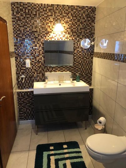 Ein modernes Badezimmer mit Mosaikfliesen und einem Waschbecken. Die Ausstattung umfasst einen Spiegel, eine Lampe und eine Toilette.