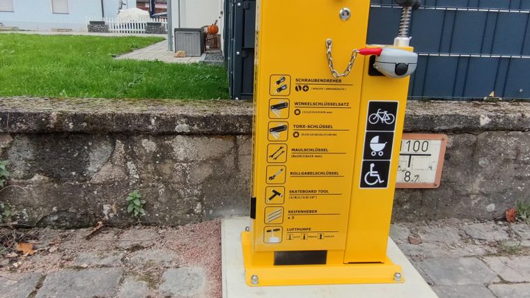 Eine gelbe Reparaturstation für Fahrräder steht an einem Gehweg. Sie enthält verschiedene Bedienungsanleitungen und ist leicht zugänglich.