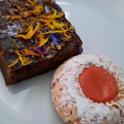 Ein Stück Schokoladenkuchen mit bunten Blütenblättern und ein Keks mit einer roten Marmeladefüllung. Beide Leckereien sind auf einem weißen Teller angerichtet.