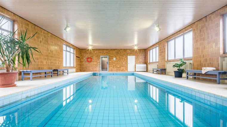 A bright indoor pool with clear water and large windows. Plants and seating areas complement the space.