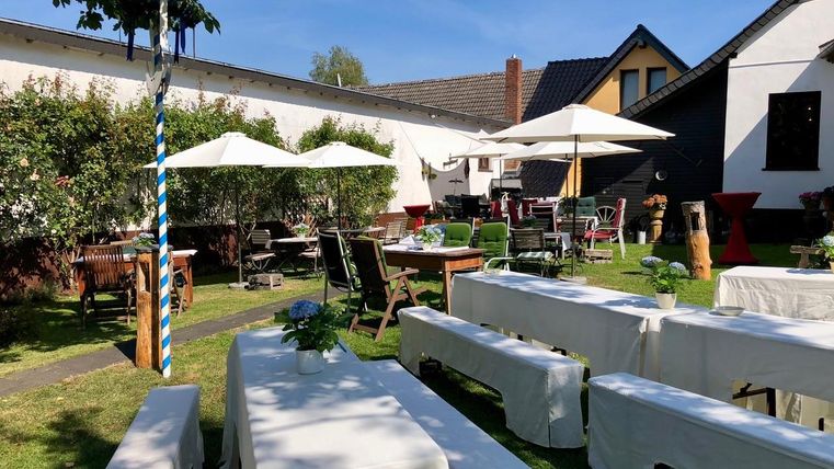 Een mooie tuin met tafels en stoelen die uitnodigen om te ontspannen. Parasols bieden schaduw, en de omgeving is groen en uitnodigend.