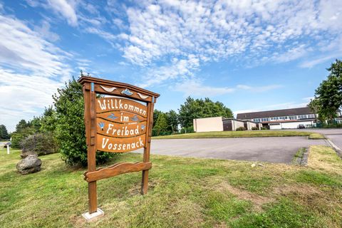 Ein Willkommensschild für das Freibad Vossenack steht auf einer Wiese. Im Hintergrund sieht man ein Gebäude und den blauen Himmel.