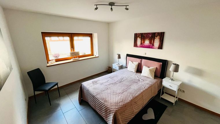 A cozy bedroom with a double bed, nightstands, and a chair. The walls are light, and a large window provides plenty of light.