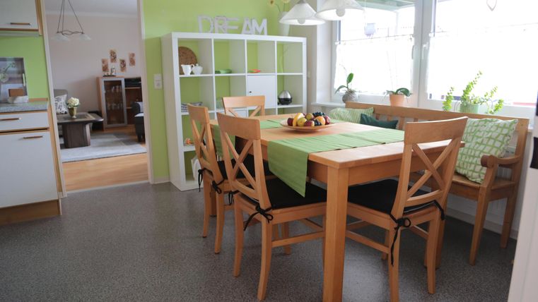 Ein helles Esszimmer mit einem großen Holztisch und Stühlen. Die Wände sind grün, und es gibt dekorative Pflanzen sowie eine offene Verbindung zum Wohnbereich.